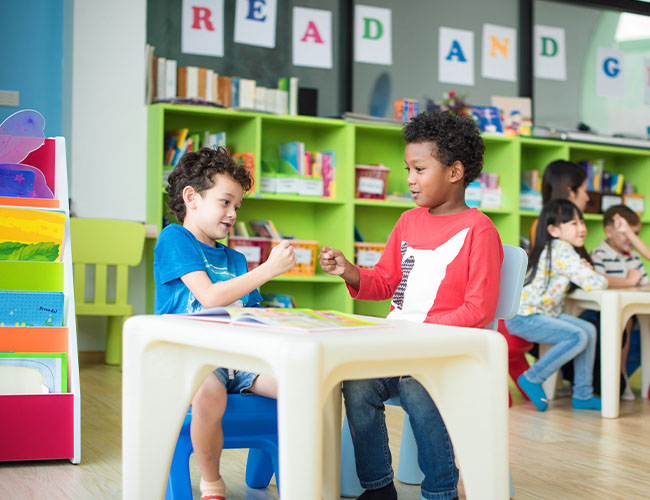 Two 4 - 5 year old kids talking at a table in a classroom.